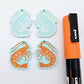 Two pairs of pastel aqua acrylic prawn dangle earring blanks with engraved details laying flat with an orange posca paint pen