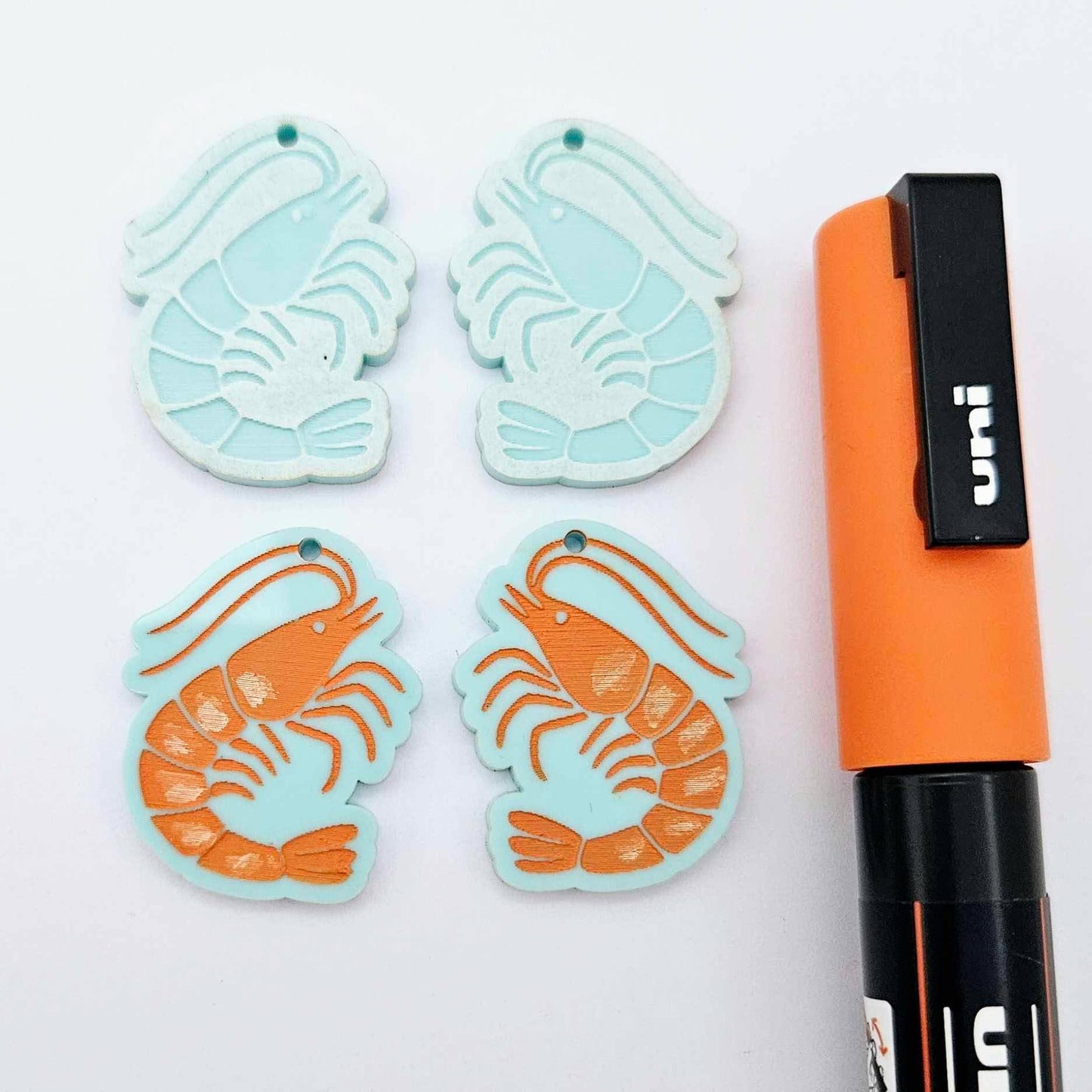 Two pairs of pastel aqua acrylic prawn dangle earring blanks with engraved details laying flat with an orange posca paint pen