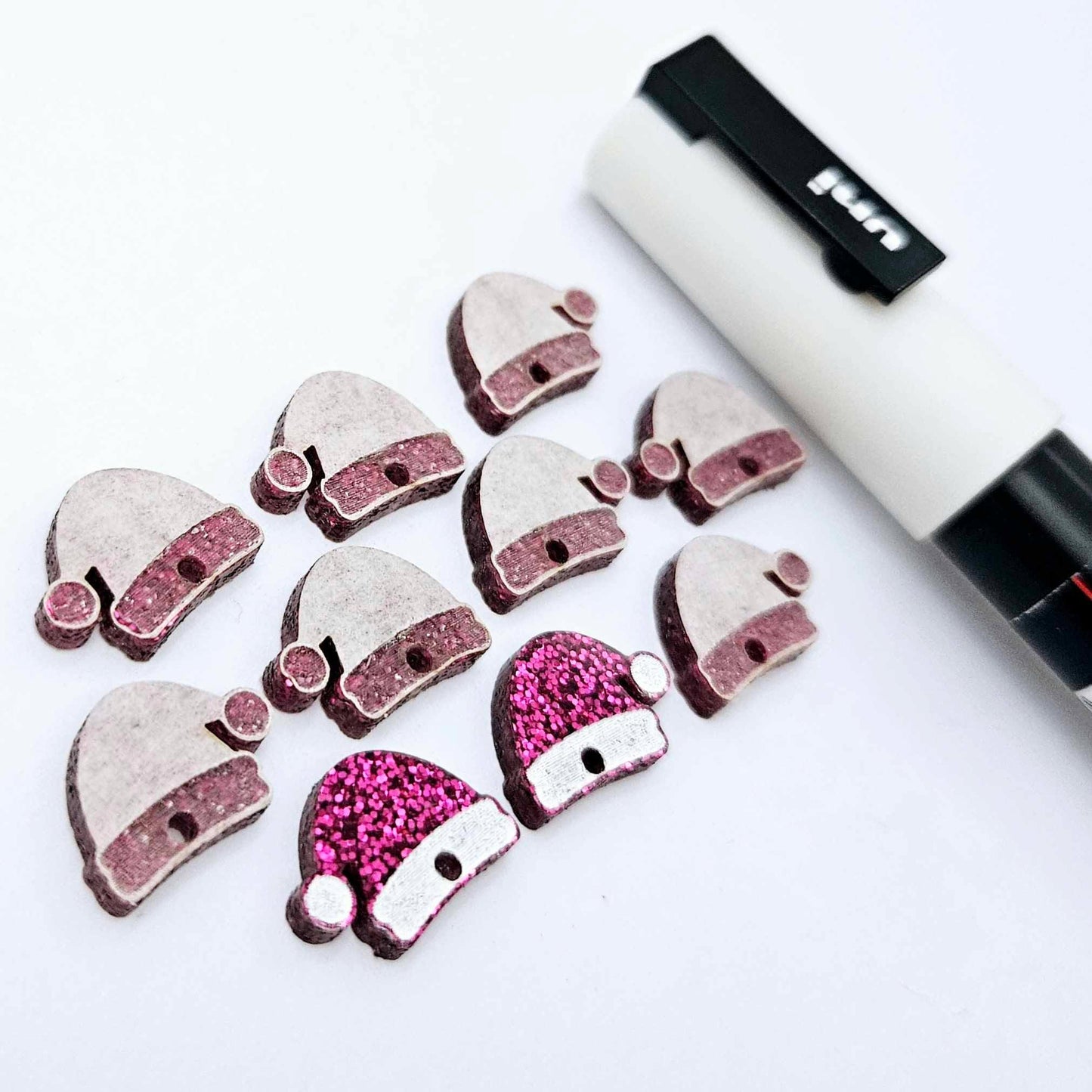 One paint-filled pair of hot pink glitter acrylic Santa hat toppers alongside multiple unpainted pieces still covered with protective paper, pictured with a White Posca Paint Pen