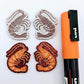 One pair of red glitter acrylic engraved prawn dangle earring blanks with protective paper on alongside a paint filled pair, laying flat with an orange posca paint pen