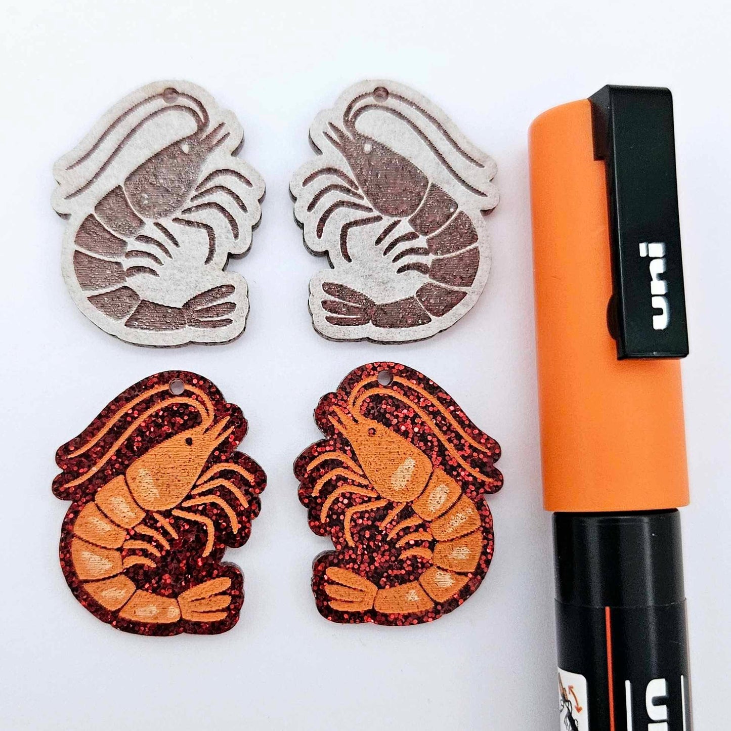 One pair of red glitter acrylic engraved prawn dangle earring blanks with protective paper on alongside a paint filled pair, laying flat with an orange posca paint pen