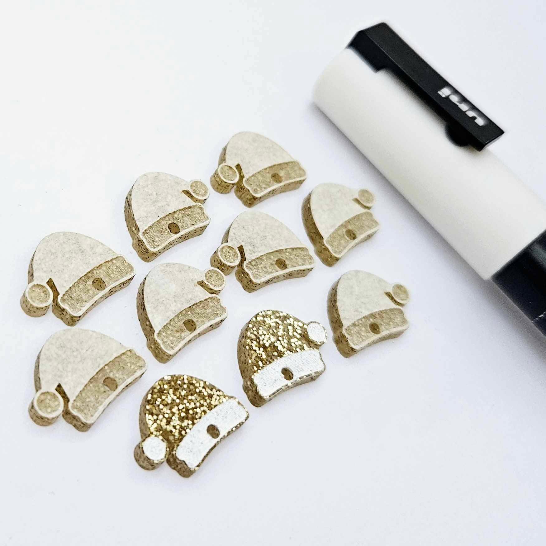One paint-filled pair of gold glitter acrylic Santa hat toppers alongside multiple unpainted pieces still covered with protective paper, pictured with a White Posca Paint Pen