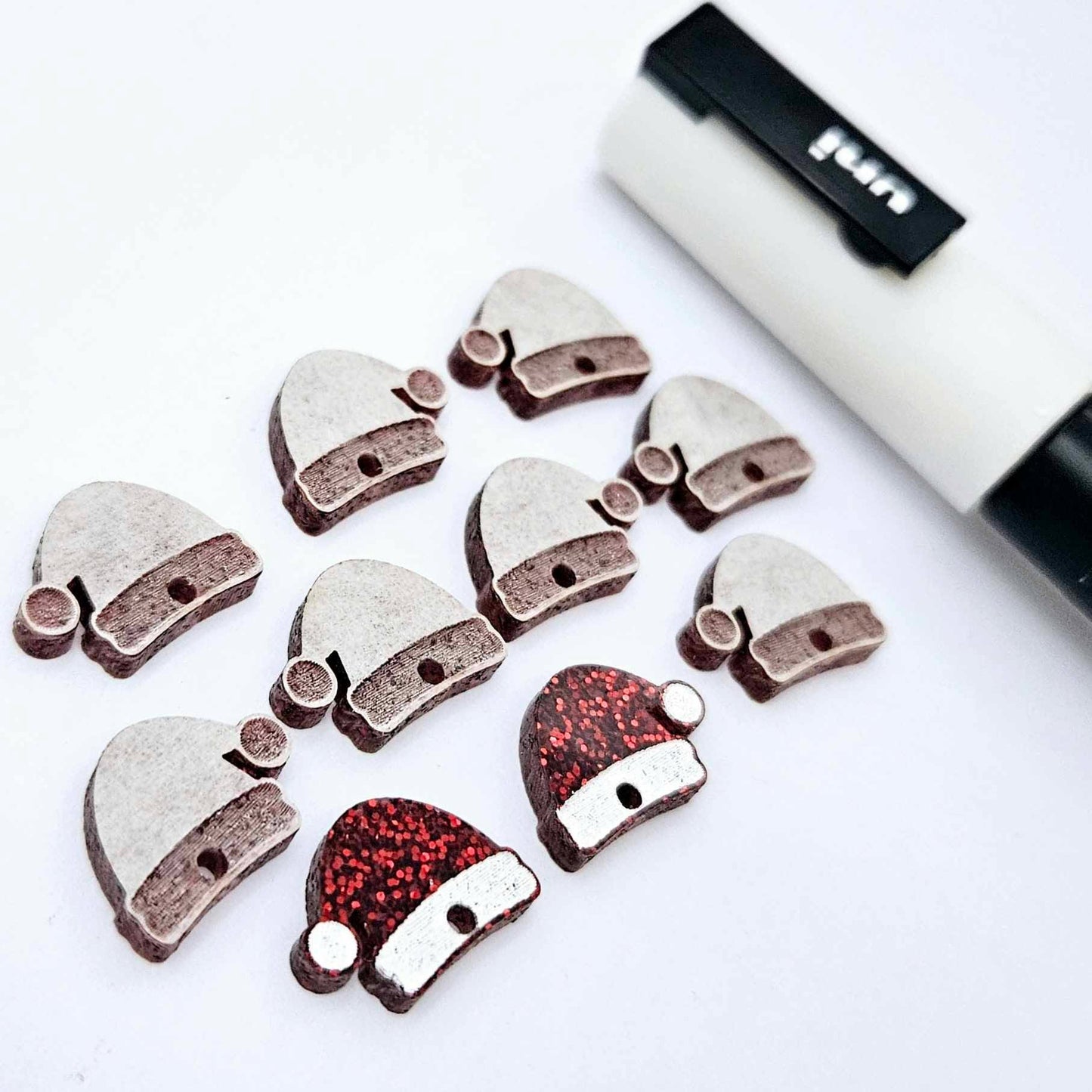 One paint-filled pair of red glitter Santa hat toppers alongside multiple unpainted pieces still covered with protective paper, pictured with a White Posca Paint Pen