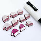 One paint-filled pair of hot pink glitter acrylic Santa hat stud blanks alongside multiple unpainted pieces still covered with protective paper, pictured with a White Posca Paint Pen