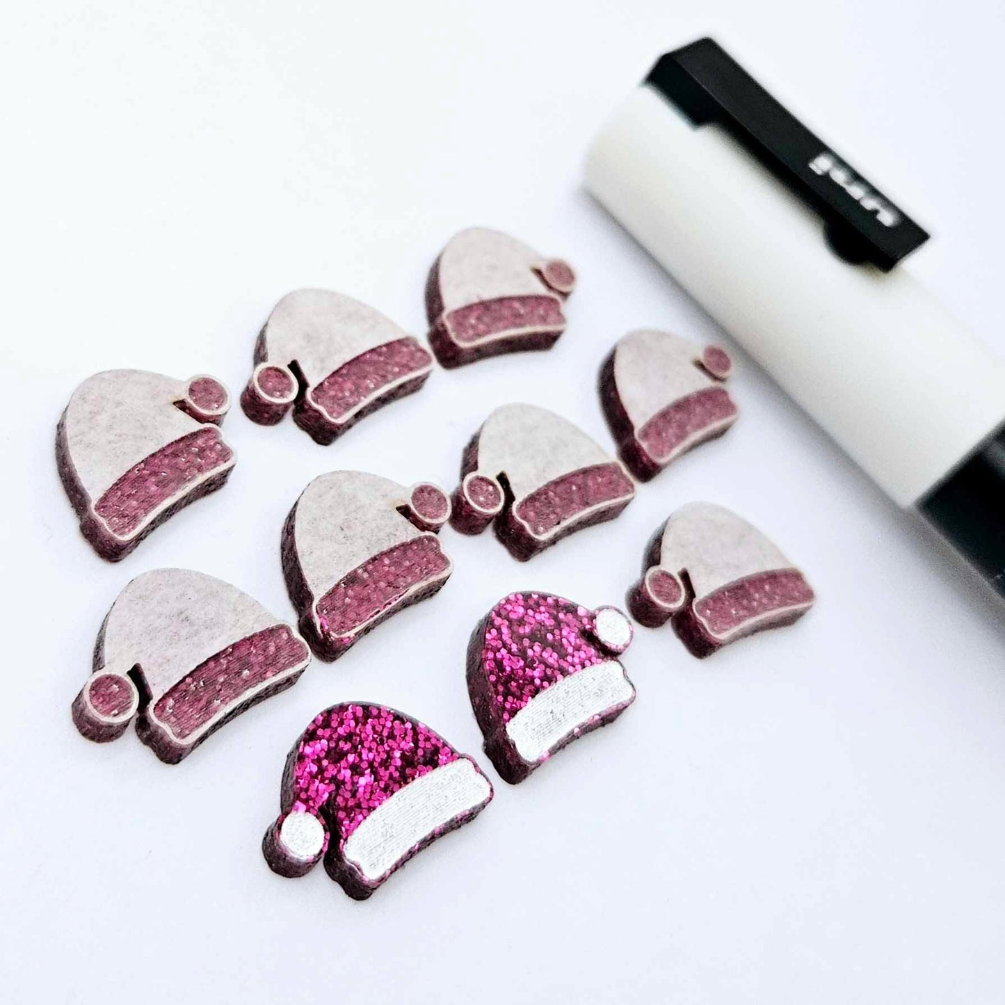 One paint-filled pair of hot pink glitter acrylic Santa hat stud blanks alongside multiple unpainted pieces still covered with protective paper, pictured with a White Posca Paint Pen