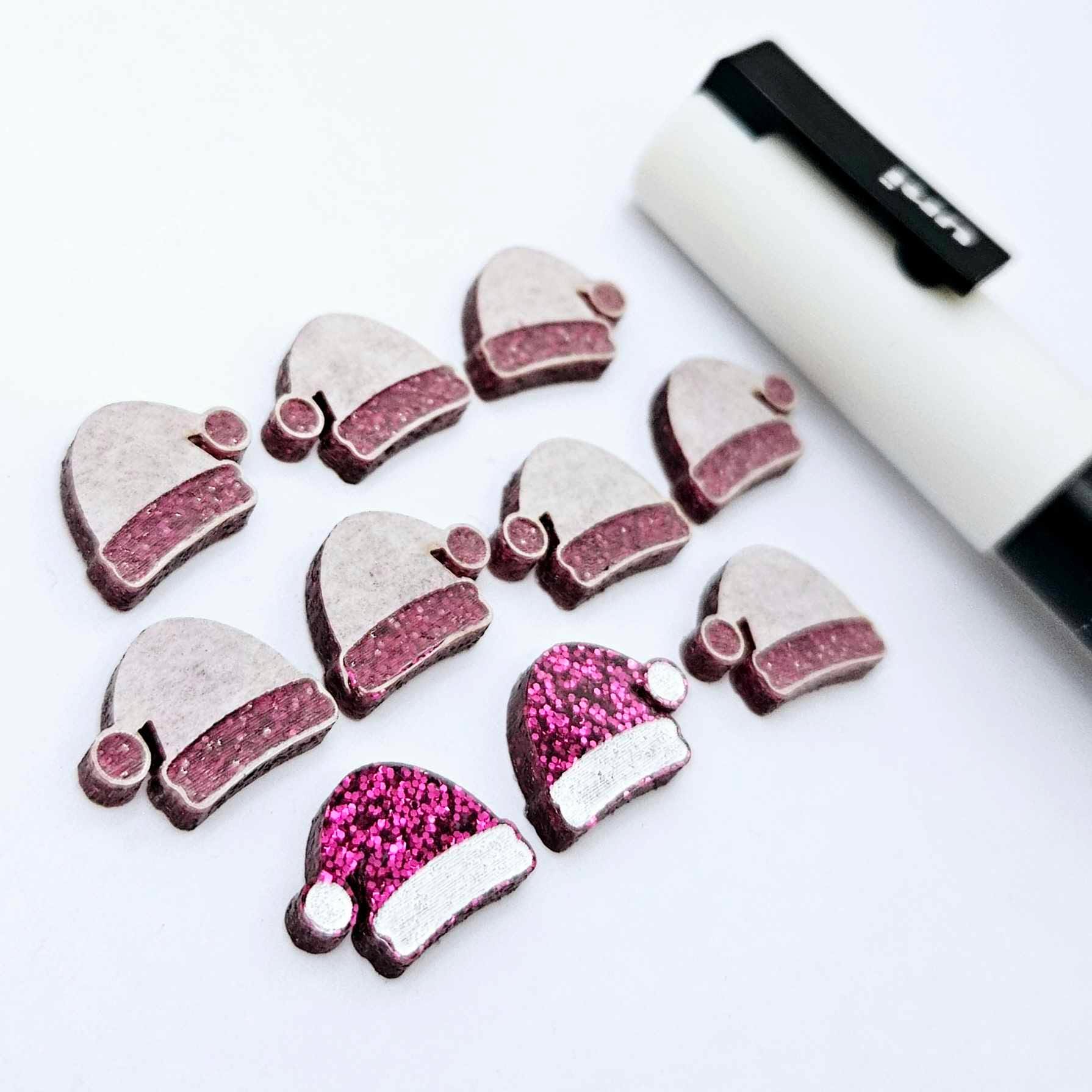 One paint-filled pair of hot pink glitter acrylic Santa hat stud blanks alongside multiple unpainted pieces still covered with protective paper, pictured with a White Posca Paint Pen