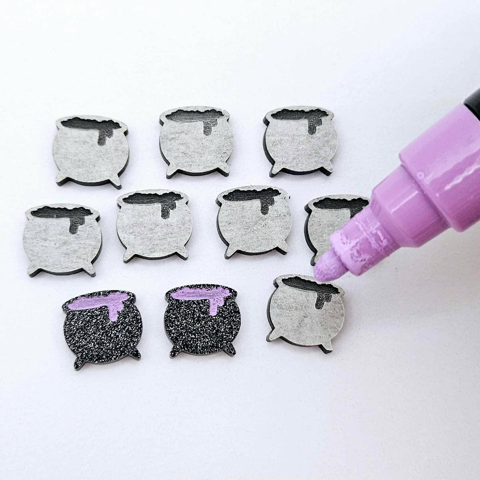 One pair of black glitter acrylic Cauldron stud earring blanks with lavender paint-filled details, surrounded by multiple unpainted pieces with protective paper on, pictured with an uncapped Lavender Posca Paint Pen ready for paint filling