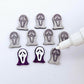 One pair of holographic purple glitter acrylic spooky face mask stud earring blanks with white paint-filled details, surrounded by multiple unpainted pieces with protective paper on, pictured with an uncapped White Posca Paint Pen ready for paint filling