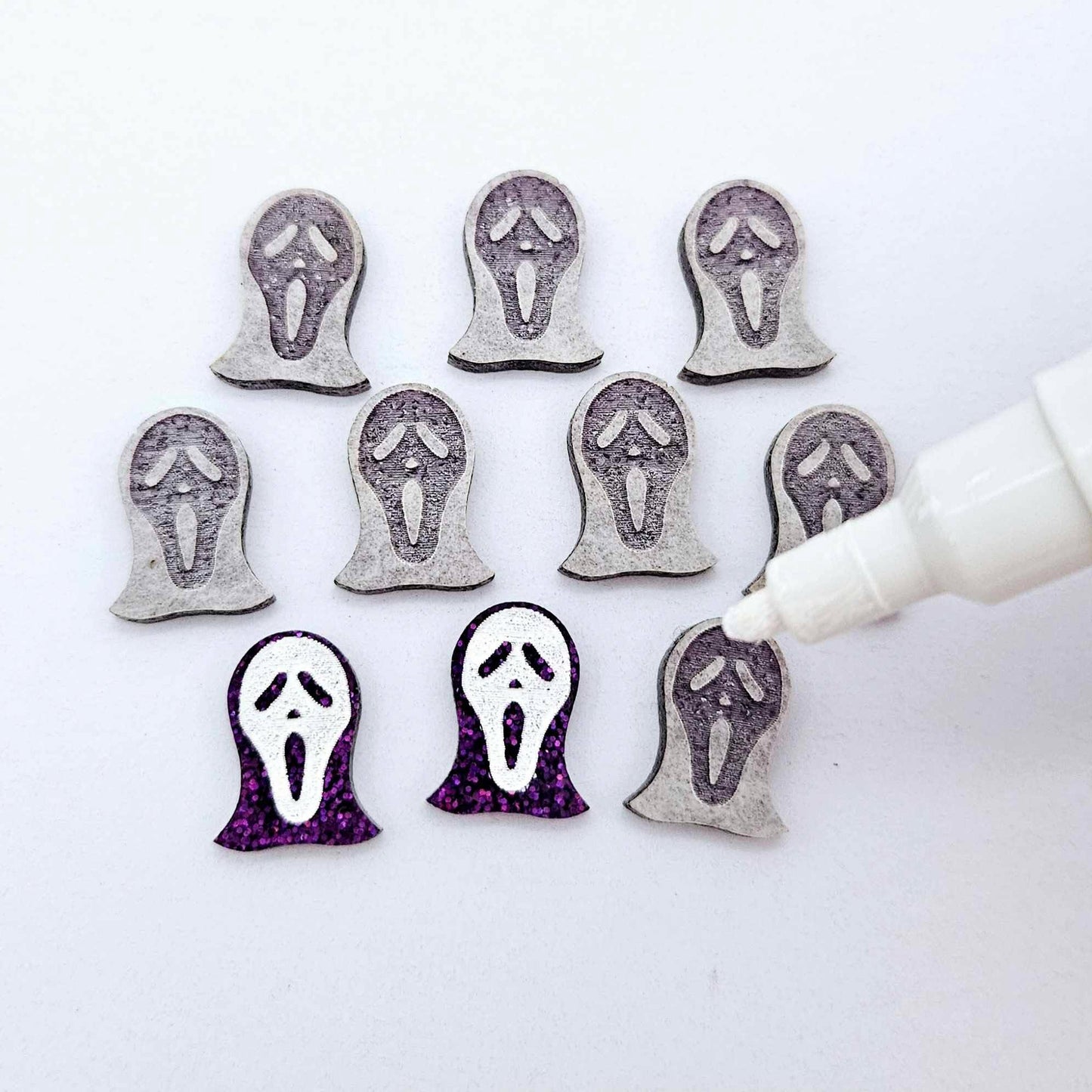 One pair of holographic purple glitter acrylic spooky face mask stud earring blanks with white paint-filled details, surrounded by multiple unpainted pieces with protective paper on, pictured with an uncapped White Posca Paint Pen ready for paint filling
