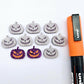 One pair of holographic purple glitter Jack-O’-Lantern stud earring blanks with orange paint-filled details, surrounded by multiple unpainted pieces with protective paper on, pictured with an Orange Posca Paint Pen