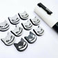 One pair of black glitter acrylic Spooky Cat stud earring blanks with white paint-filled details, surrounded by multiple unpainted pieces with protective paper on, pictured with a White Posca Paint Pen