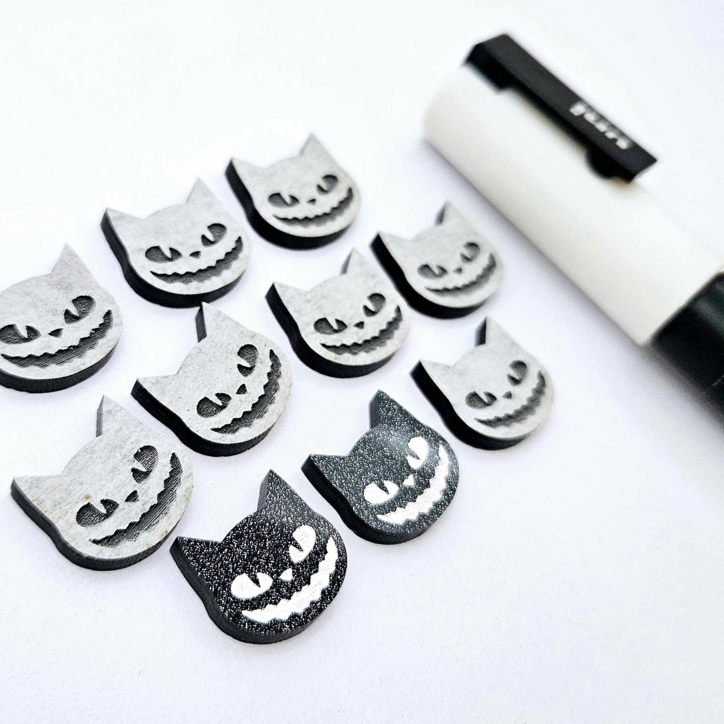 One pair of black glitter acrylic Spooky Cat stud earring blanks with white paint-filled details, surrounded by multiple unpainted pieces with protective paper on, pictured with a White Posca Paint Pen