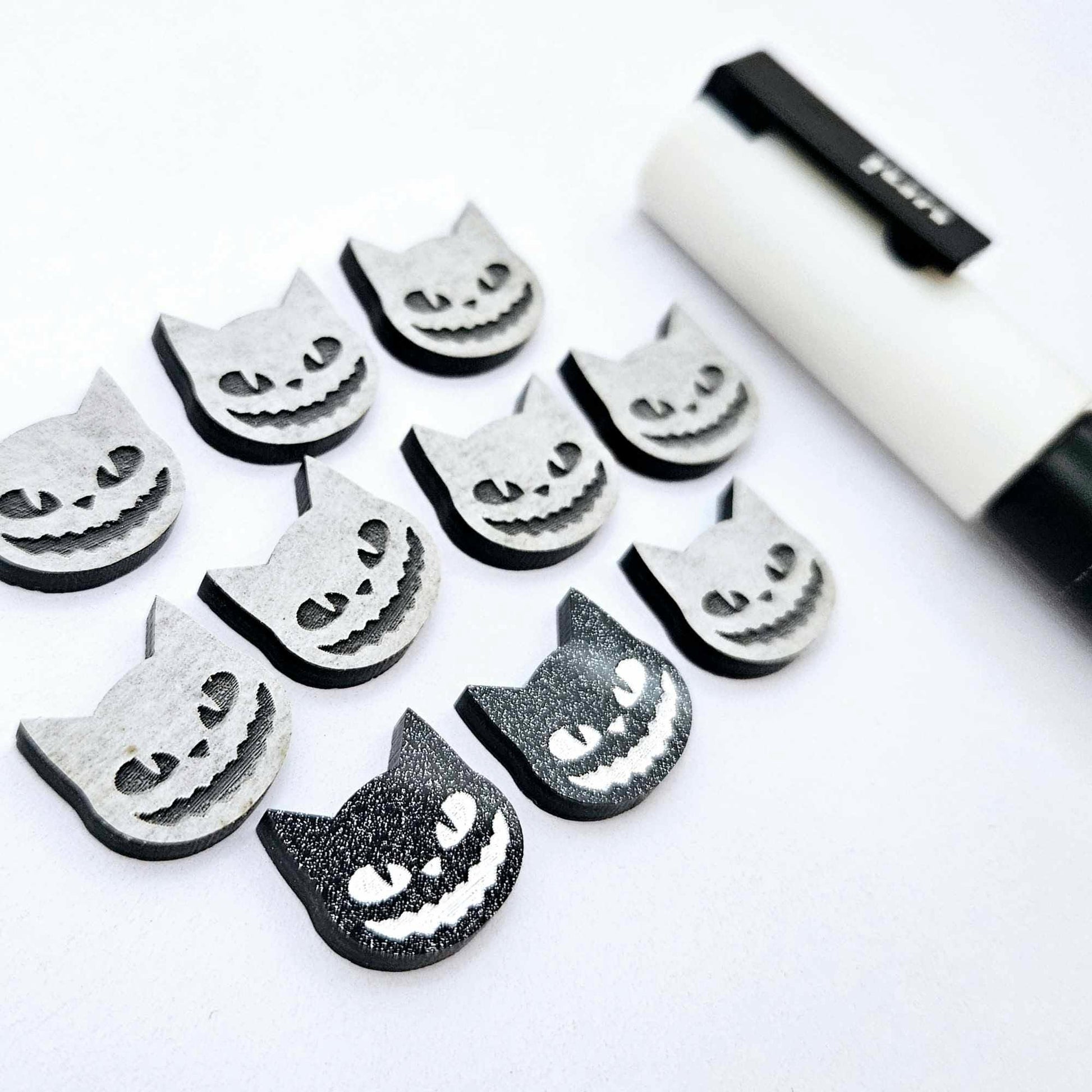 One pair of black glitter acrylic Spooky Cat stud earring blanks with white paint-filled details, surrounded by multiple unpainted pieces with protective paper on, pictured with a White Posca Paint Pen