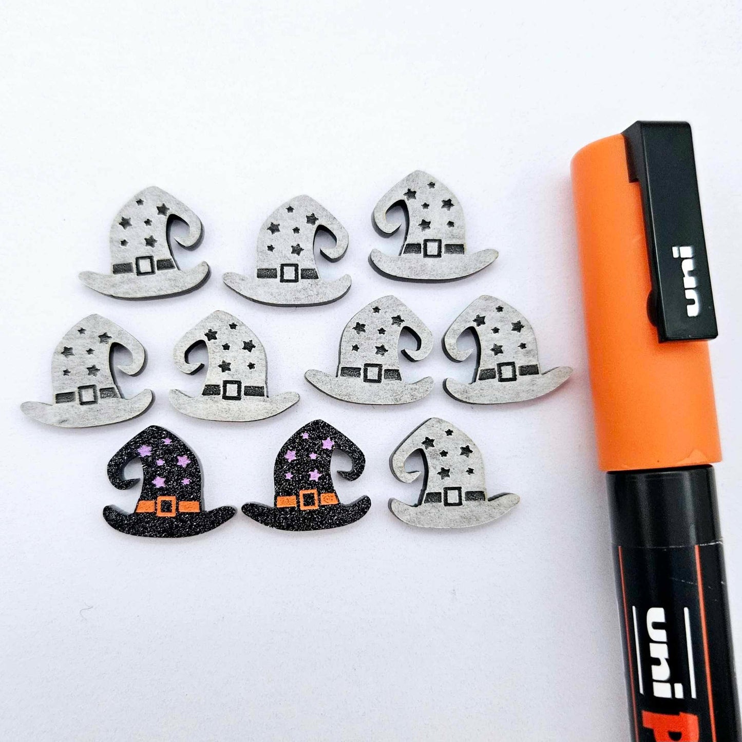 One pair of black glitter acrylic witch’s hat stud earring blanks with orange and lavender paint-filled details, surrounded by multiple unpainted pieces with protective paper on, pictured with an orange Posca Paint Pen