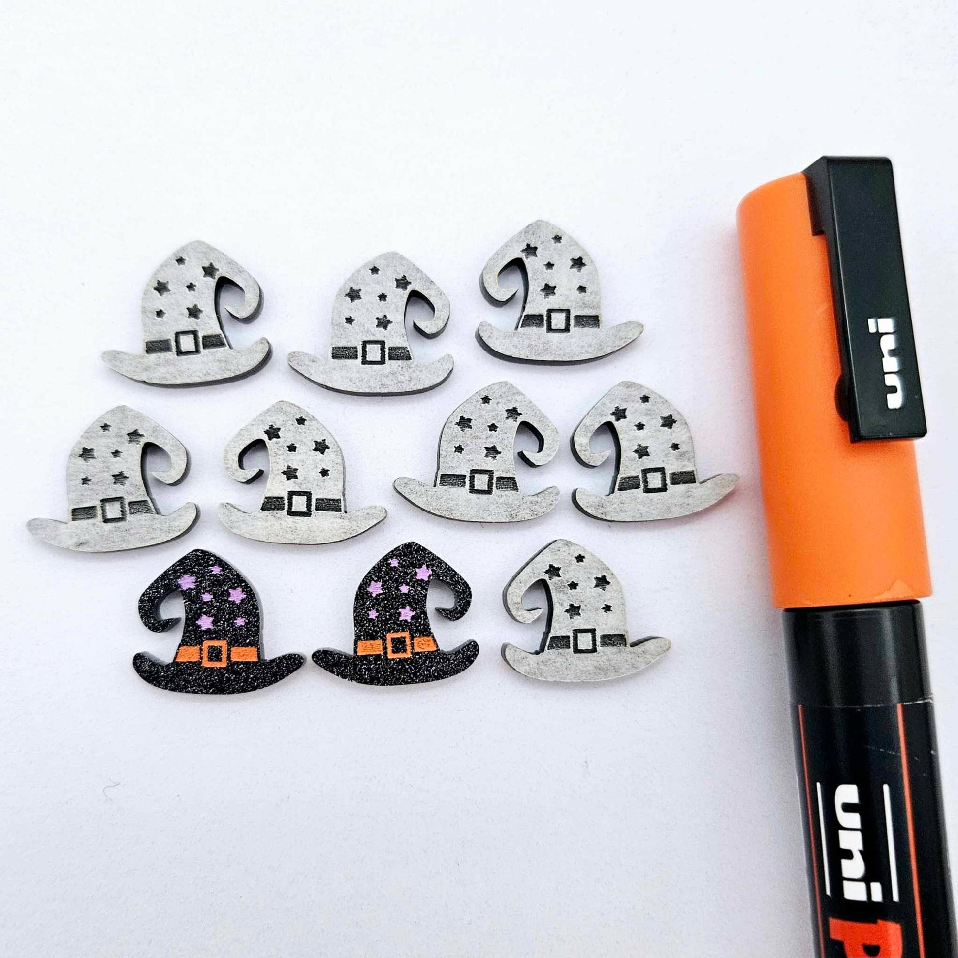 One pair of black glitter acrylic witch’s hat stud earring blanks with orange and lavender paint-filled details, surrounded by multiple unpainted pieces with protective paper on, pictured with an orange Posca Paint Pen