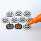 One pair of black glitter acrylic Jack-O’-Lantern stud earring blanks with orange paint-filled details, surrounded by multiple unpainted pieces with protective paper on, pictured with an uncapped Orange Posca Paint Pen ready for paint filling