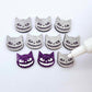 One pair of holographic purple glitter acrylic Spooky Cat stud earring blanks with white paint-filled details, surrounded by multiple unpainted pieces with protective paper on, pictured with an uncapped White Posca Paint Pen ready for paint filling