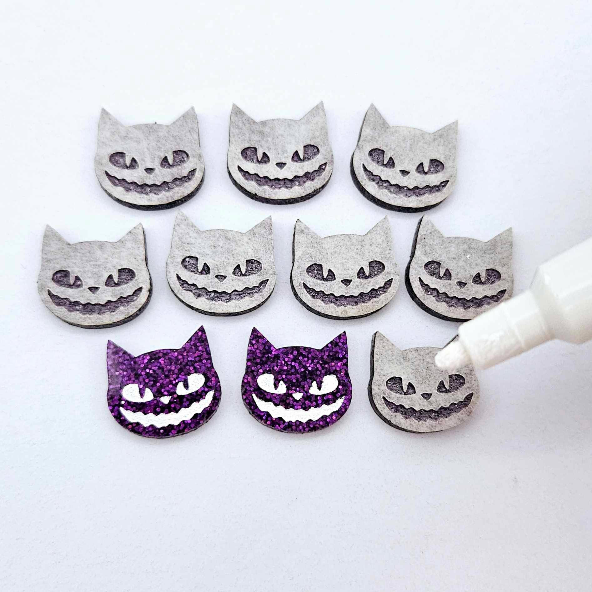 One pair of holographic purple glitter acrylic Spooky Cat stud earring blanks with white paint-filled details, surrounded by multiple unpainted pieces with protective paper on, pictured with an uncapped White Posca Paint Pen ready for paint filling