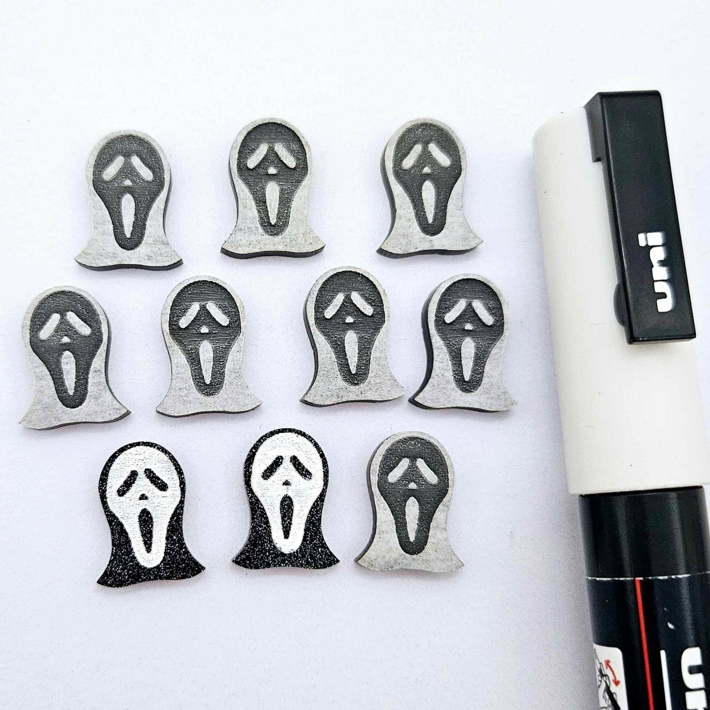 One pair of black glitter acrylic spooky face mask stud earring blanks with white paint-filled details, surrounded by multiple unpainted pieces with protective paper on, pictured with a White Posca Paint Pen