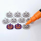 One pair of holographic purple glitter acrylic Jack-O’-Lantern stud earring blanks with orange paint-filled details, surrounded by multiple unpainted pieces with protective paper on, pictured with an uncapped Orange Posca Paint Pen ready for paint filling
