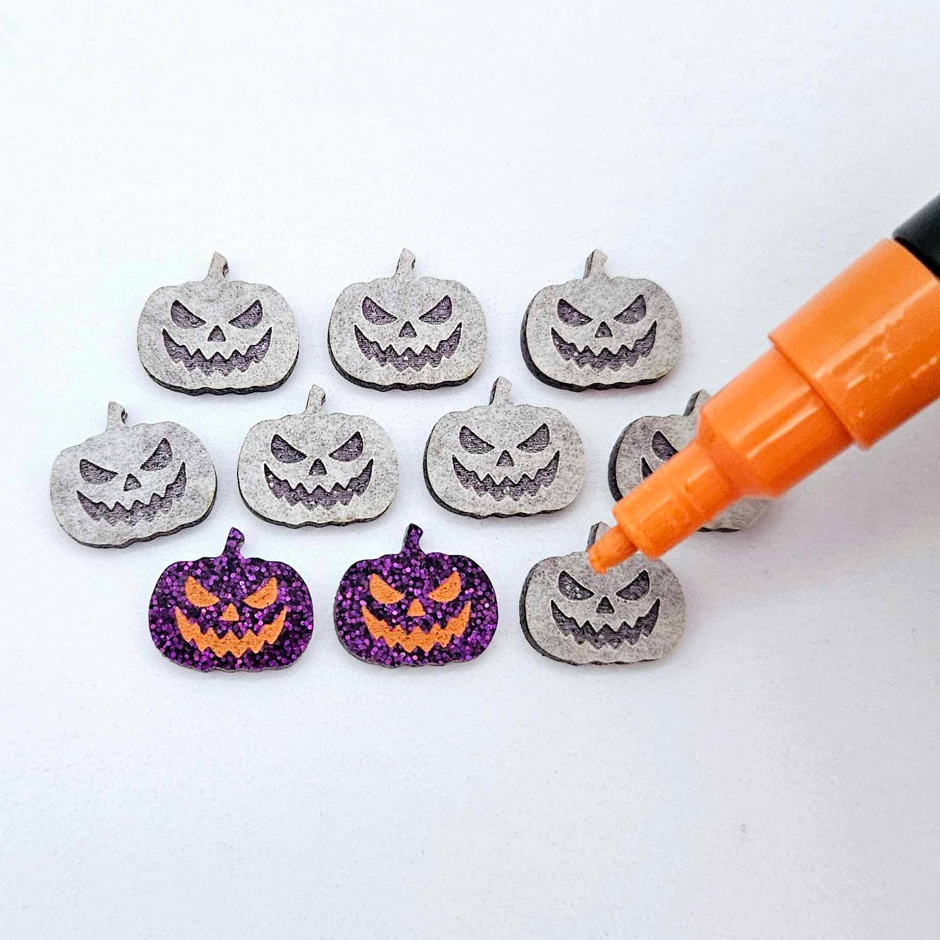One pair of holographic purple glitter acrylic Jack-O’-Lantern stud earring blanks with orange paint-filled details, surrounded by multiple unpainted pieces with protective paper on, pictured with an uncapped Orange Posca Paint Pen ready for paint filling