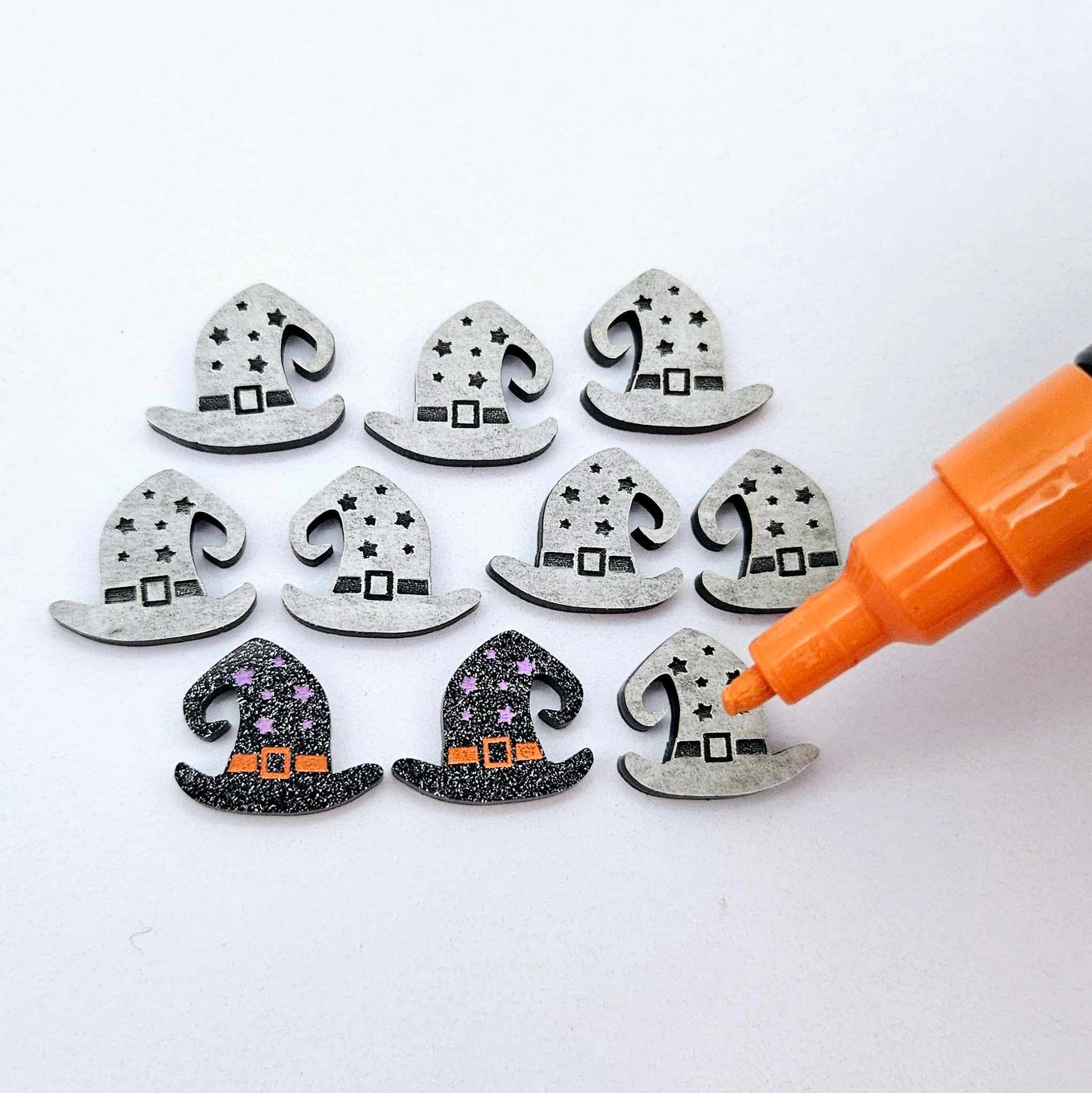 One pair of black glitter acrylic witch’s hat stud earring blanks with orange and lavender paint-filled details, surrounded by multiple unpainted pieces with protective paper on, pictured with an uncapped orange Posca Paint Pen ready for paint filling