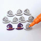 One pair of holographic purple glitter acrylic witch’s hat stud earring blanks with orange and green paint-filled details, surrounded by multiple unpainted pieces with protective paper on, pictured with an uncapped Orange Posca Paint Pen ready for paint filling