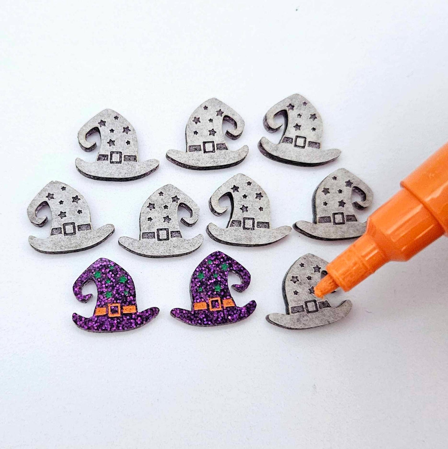 One pair of holographic purple glitter acrylic witch’s hat stud earring blanks with orange and green paint-filled details, surrounded by multiple unpainted pieces with protective paper on, pictured with an uncapped Orange Posca Paint Pen ready for paint filling