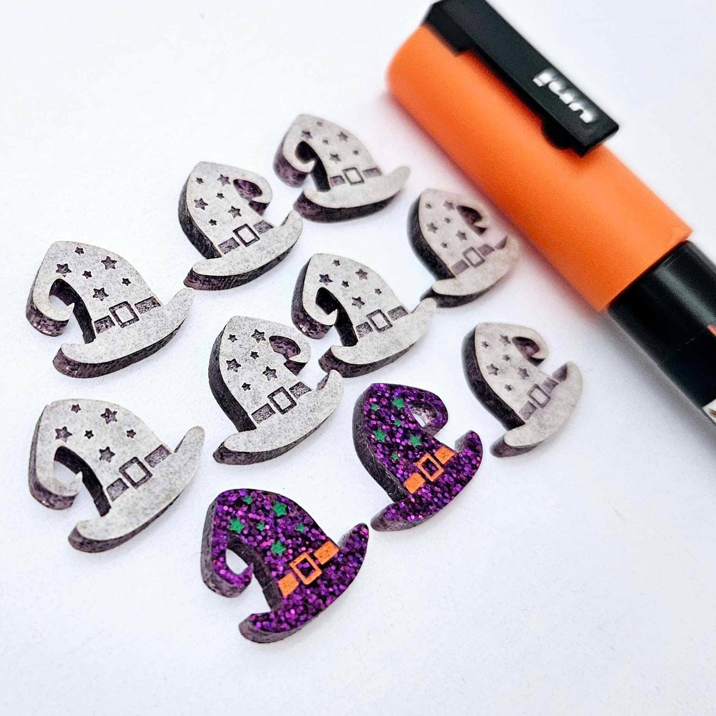 One pair of holographic purple glitter acrylic witch’s hat stud earring blanks with orange and green paint-filled details, surrounded by multiple unpainted pieces with protective paper on, pictured with an Orange Posca Paint Pen