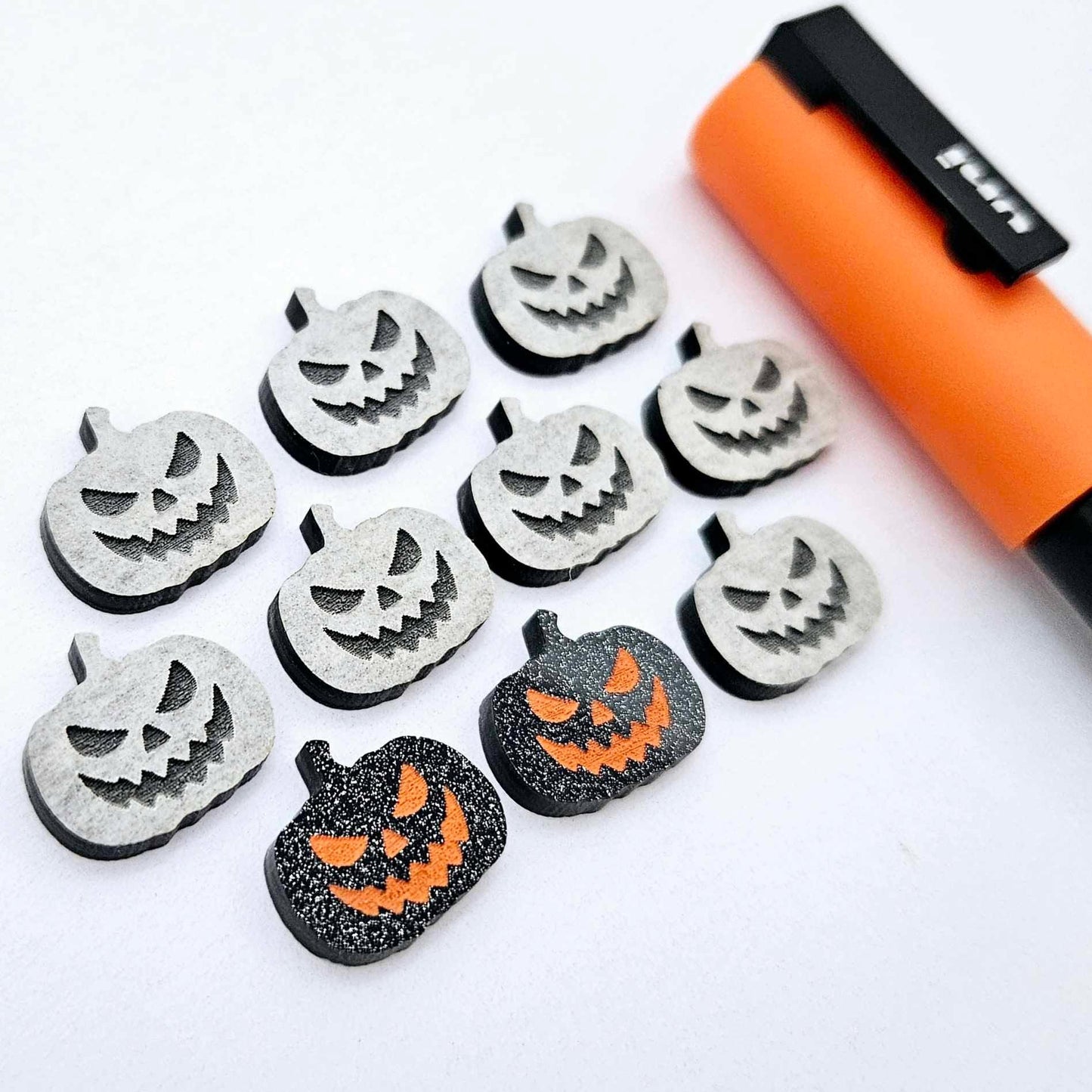 One pair of black glitter acrylic Jack-O’-Lantern stud earring blanks with orange paint-filled details, surrounded by multiple unpainted pieces with protective paper on, pictured with an Orange Posca Paint Pen
