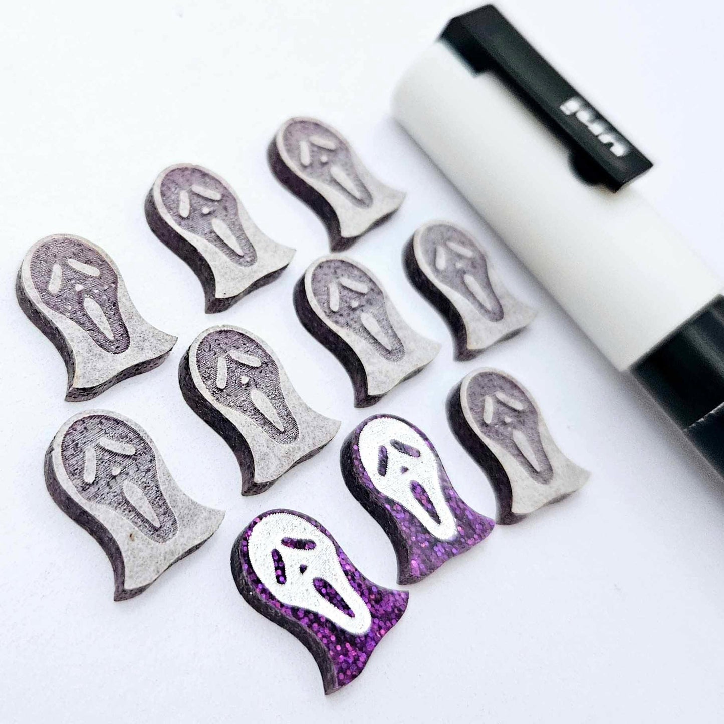 One pair of holographic purple glitter acrylic spooky face mask stud earring blanks with white paint-filled details, surrounded by multiple unpainted pieces with protective paper on, pictured with a White Posca Paint Pen