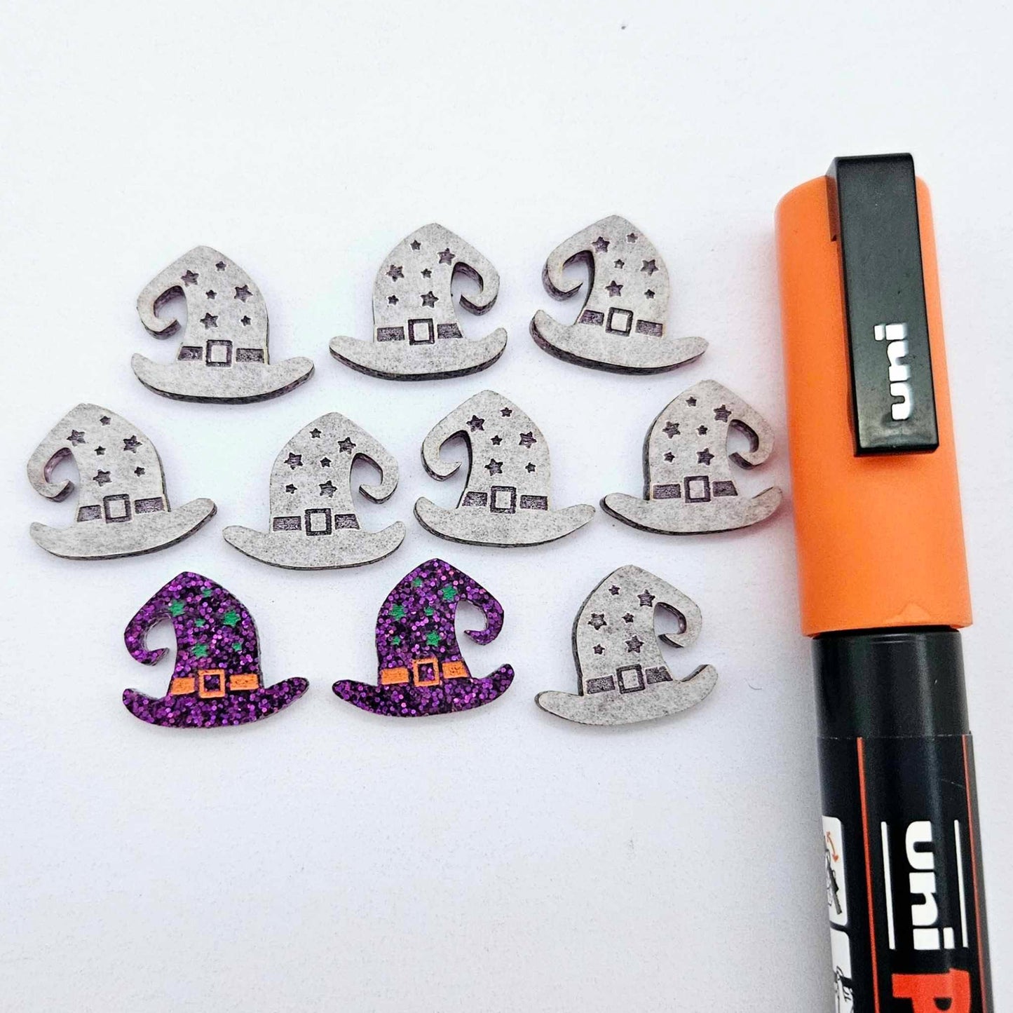 One pair of holographic purple glitter acrylic witch’s hat stud earring blanks with orange and green paint-filled details, surrounded by multiple unpainted pieces with protective paper on, pictured with an Orange Posca Paint Pen