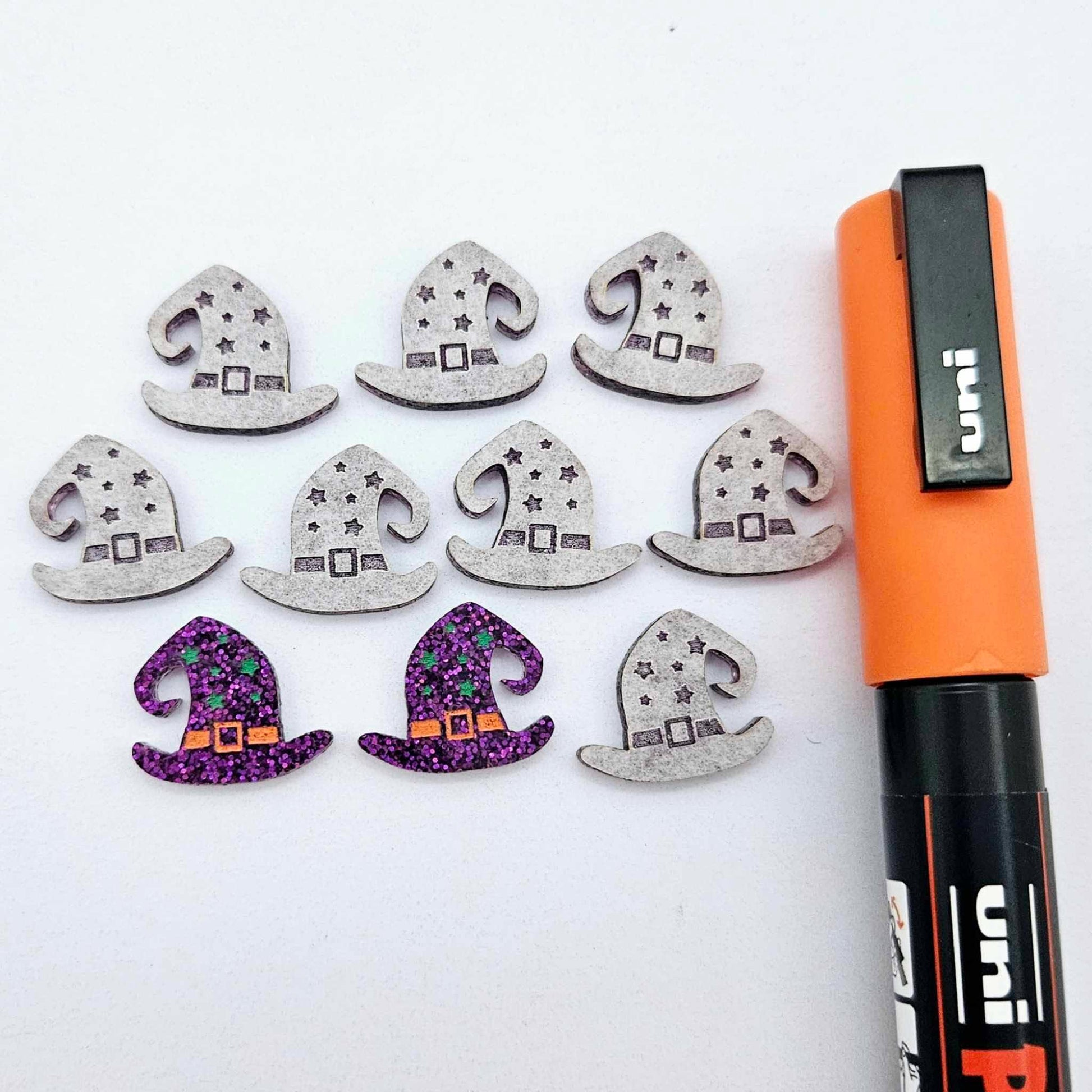 One pair of holographic purple glitter acrylic witch’s hat stud earring blanks with orange and green paint-filled details, surrounded by multiple unpainted pieces with protective paper on, pictured with an Orange Posca Paint Pen