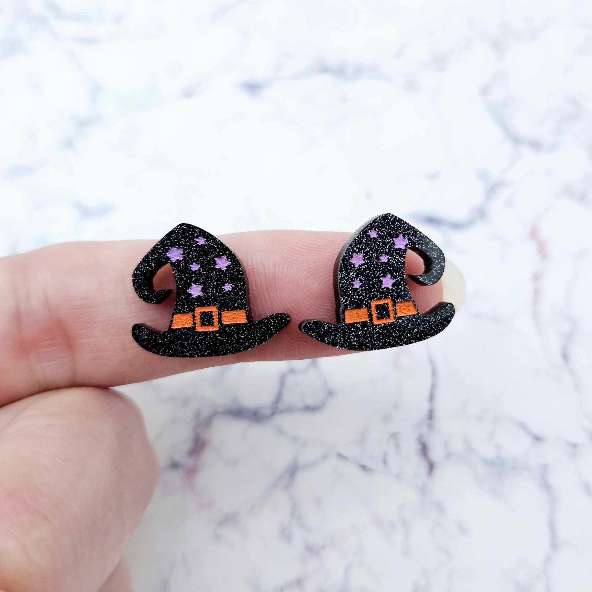 One pair of 18mm black glitter acrylic witch’s hat stud earring blanks with orange and lavender paint-filled details shown on a finger for size reference