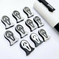 One pair of black glitter acrylic spooky face mask stud earring blanks with white paint-filled details, surrounded by multiple unpainted pieces with protective paper on, pictured with a White Posca Paint Pen