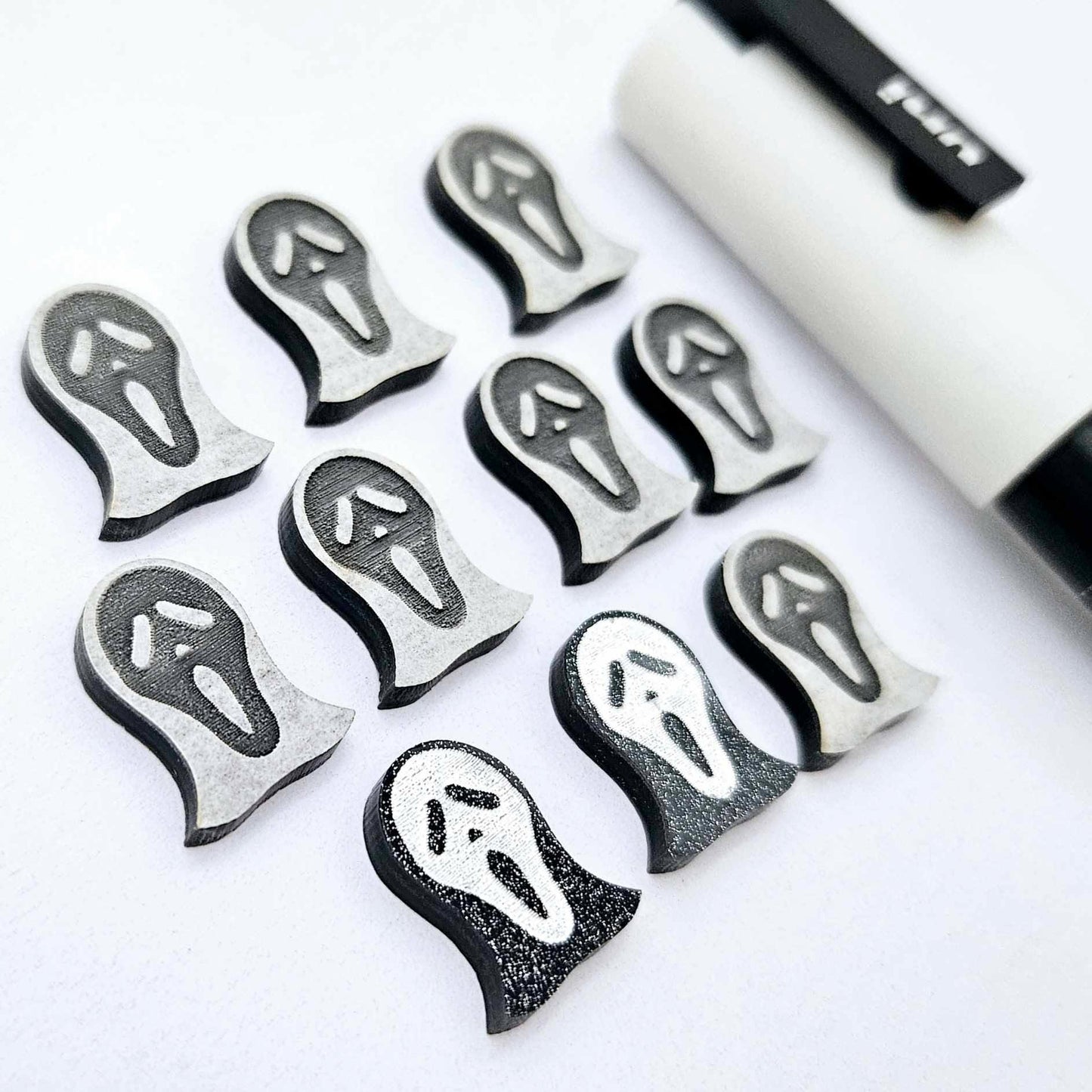 One pair of black glitter acrylic spooky face mask stud earring blanks with white paint-filled details, surrounded by multiple unpainted pieces with protective paper on, pictured with a White Posca Paint Pen