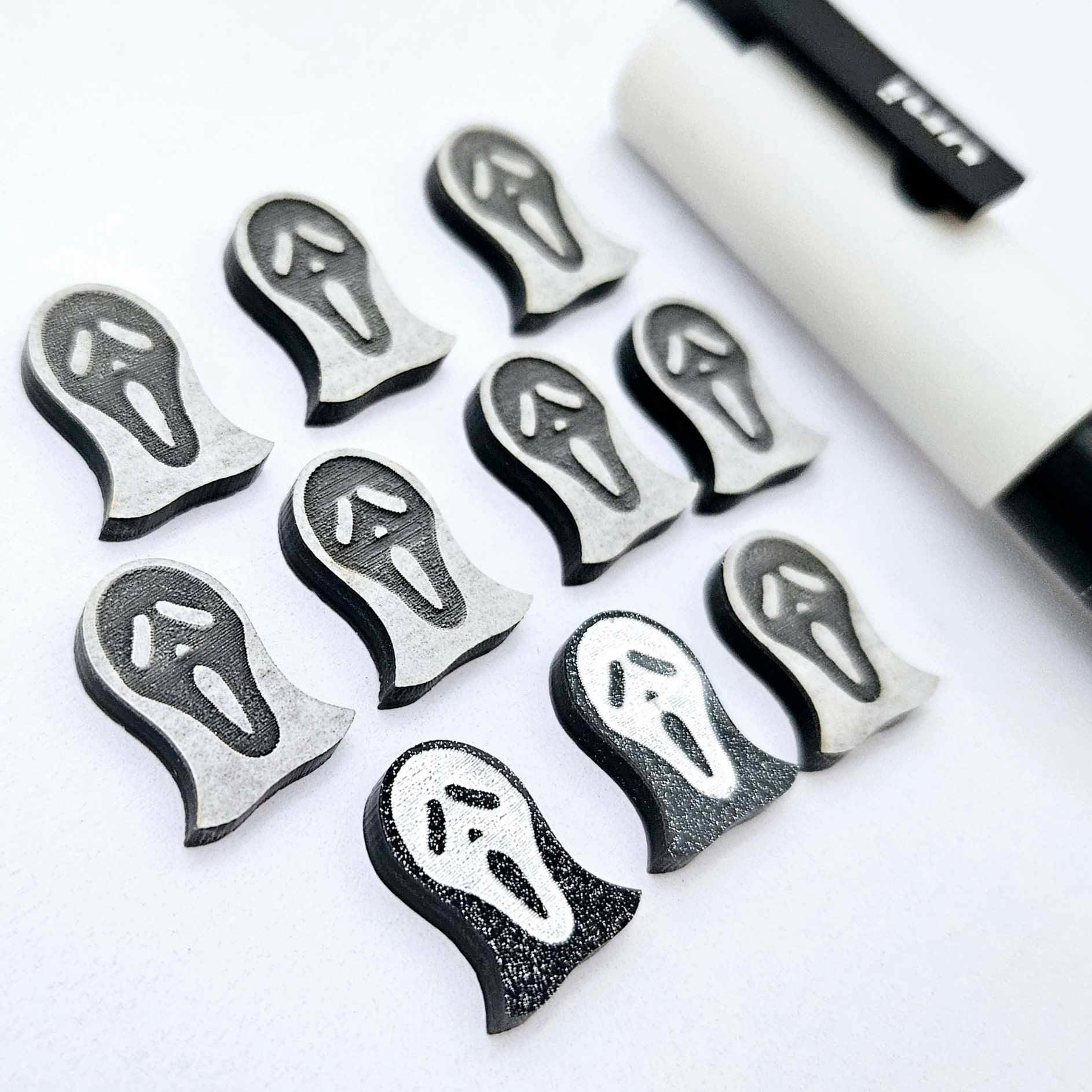One pair of black glitter acrylic spooky face mask stud earring blanks with white paint-filled details, surrounded by multiple unpainted pieces with protective paper on, pictured with a White Posca Paint Pen