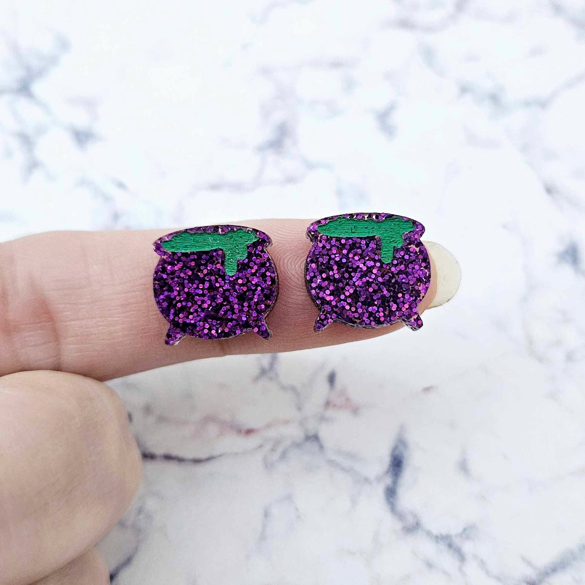 One pair of 14mm holographic purple glitter acrylic Witch's Cauldron stud earring blanks with green paint-filled bubbling potion details shown on a finger for size reference