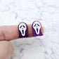 One pair of 16mm holographic purple glitter spooky face mask stud earring blanks with white paint-filled facial details shown on a finger for size reference