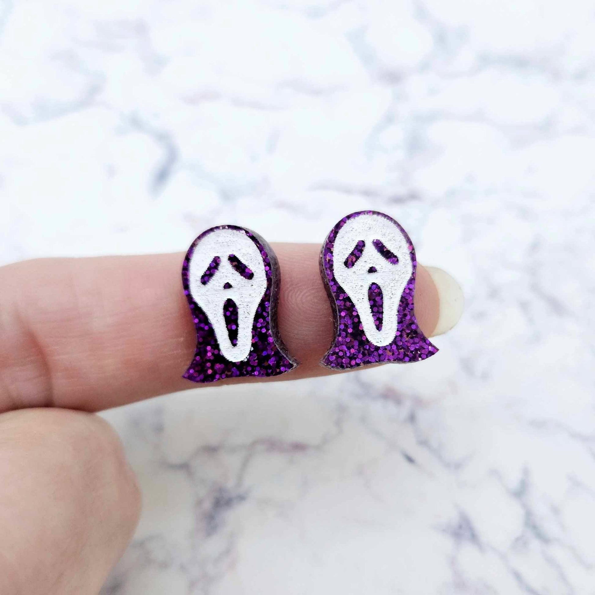One pair of 16mm holographic purple glitter spooky face mask stud earring blanks with white paint-filled facial details shown on a finger for size reference