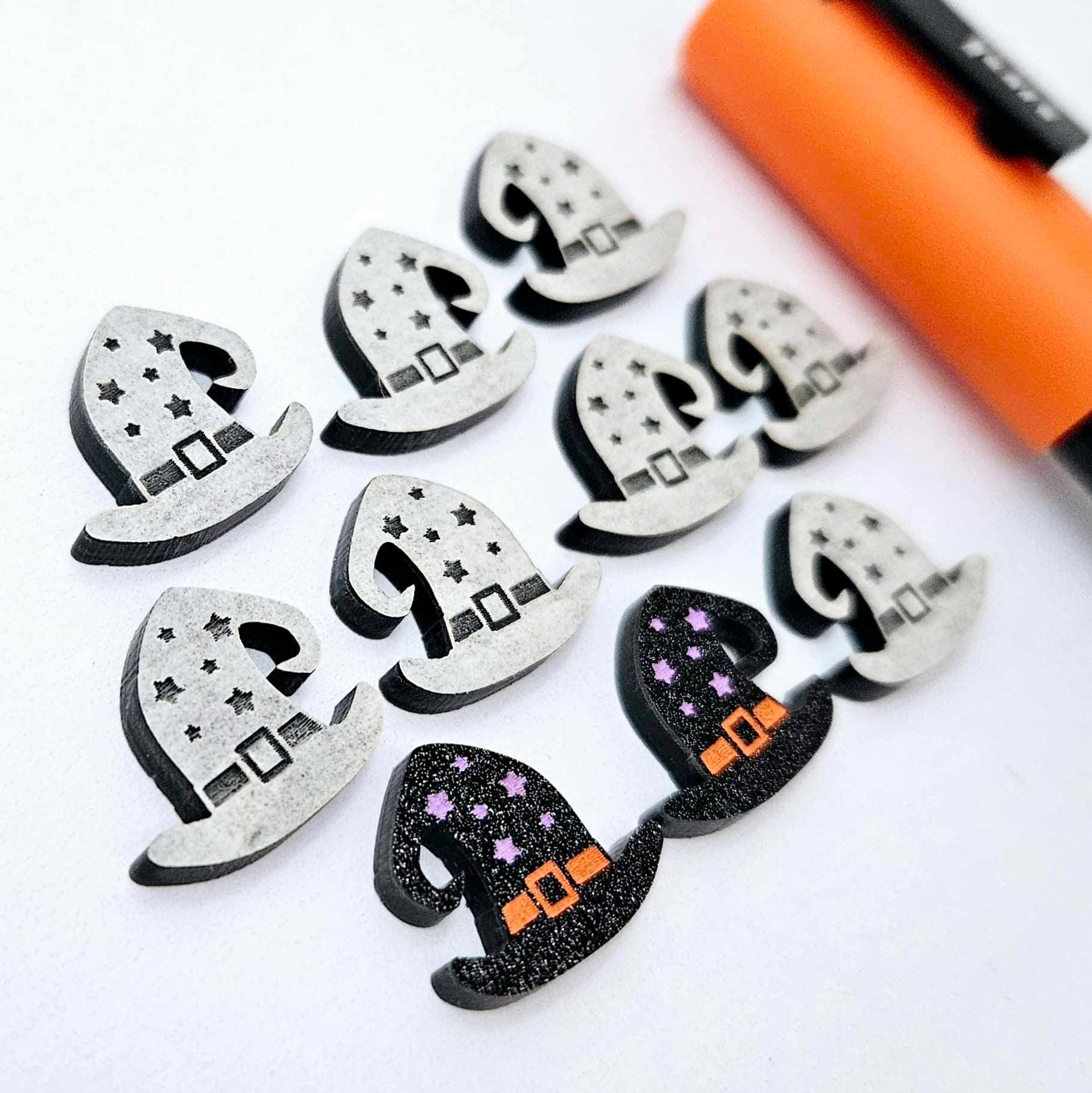 One pair of black glitter acrylic witch’s hat stud earring blanks with orange and lavender paint-filled details, surrounded by multiple unpainted pieces with protective paper on, pictured with an orange Posca Paint Pen
