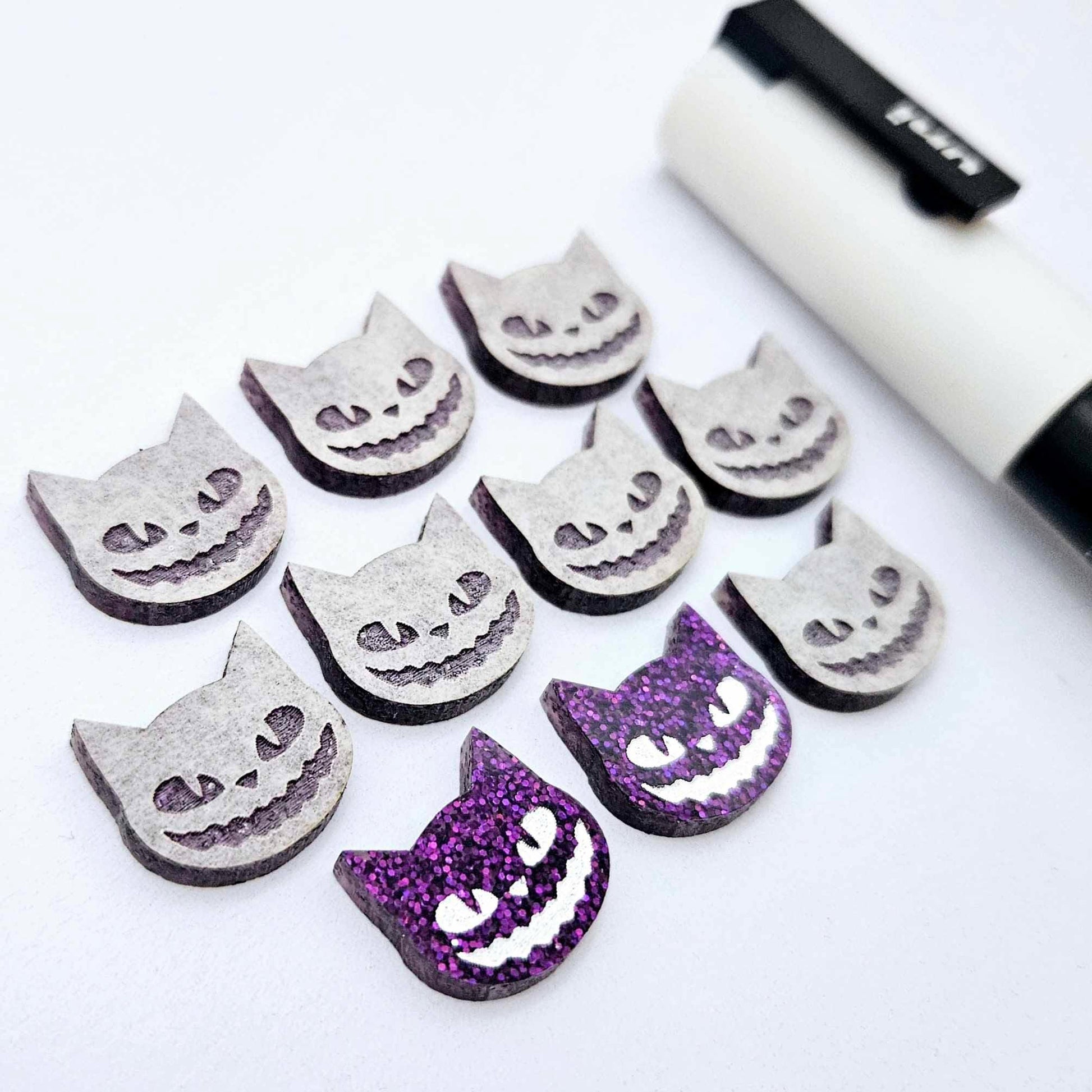 One pair of holographic purple glitter acrylic Spooky Cat stud earring blanks with white paint-filled details, surrounded by multiple unpainted pieces with protective paper on, pictured with a White Posca Paint Pen