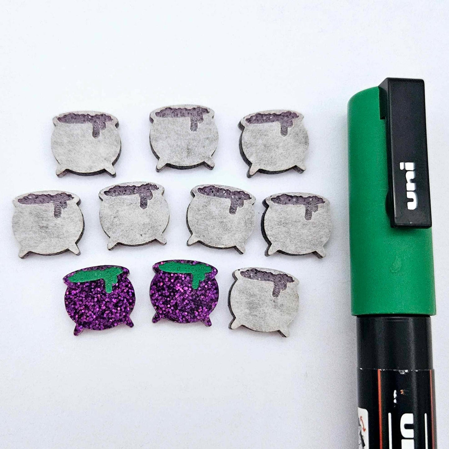 One pair of holographic purple glitter acrylic Witch's Cauldron stud earring blanks with green paint-filled details, surrounded by multiple unpainted pieces with protective paper on, pictured with a Green Posca Paint Pen