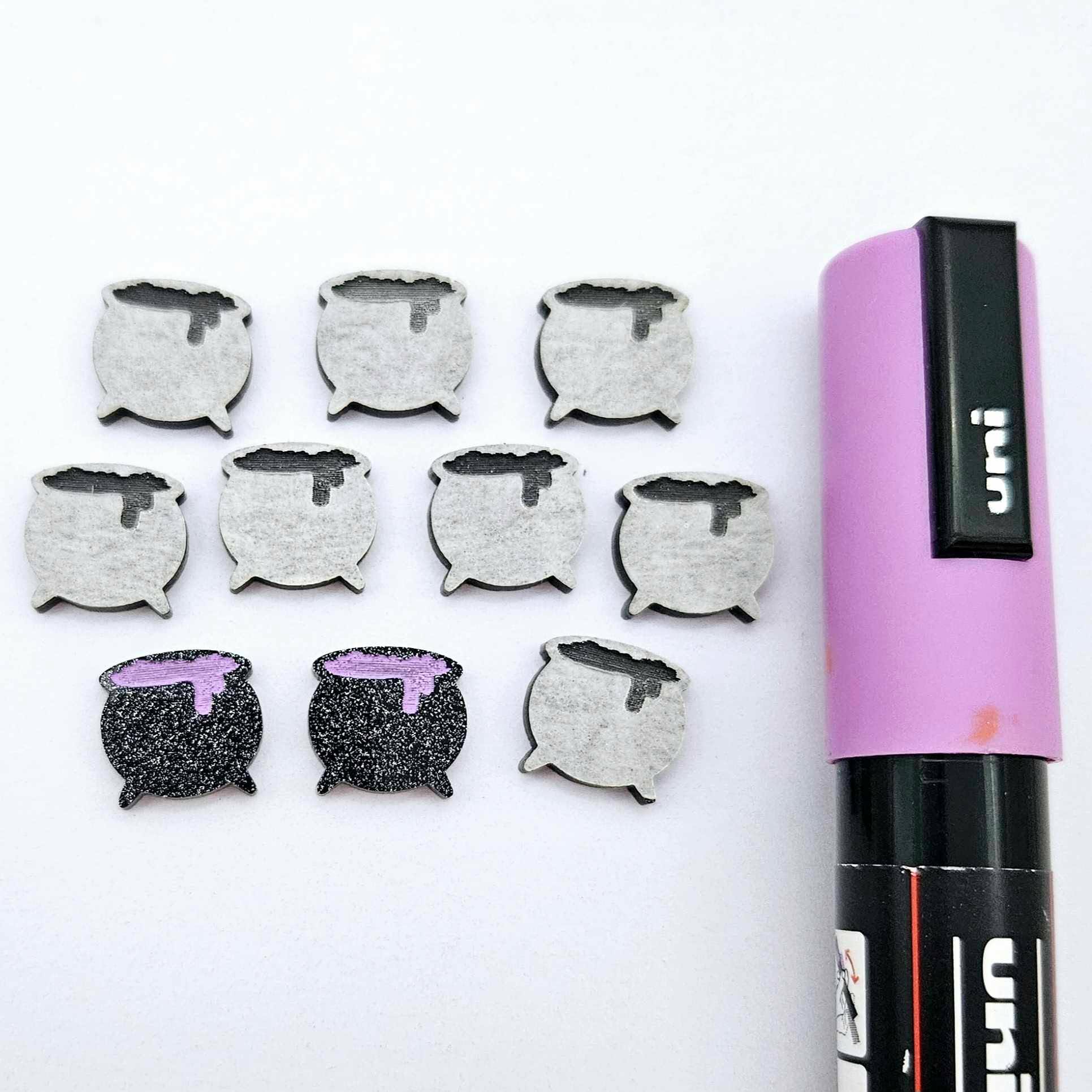 One pair of black glitter acrylic Witch's Cauldron stud earring blanks with lavender paint-filled details, surrounded by multiple unpainted pieces with protective paper on, pictured with a Lavender Posca Paint Pen.