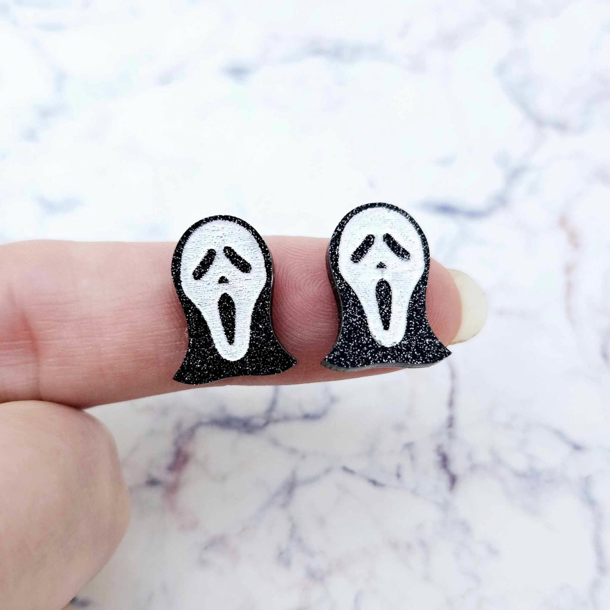 One pair of 16mm black glitter acrylic spooky face mask stud earring blanks with white paint-filled facial details shown on a finger for size reference