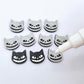 One pair of black glitter acrylic Spooky Cat stud earring blanks with white paint-filled details, surrounded by multiple unpainted pieces with protective paper on, pictured with an uncapped White Posca Paint Pen ready for paint filling