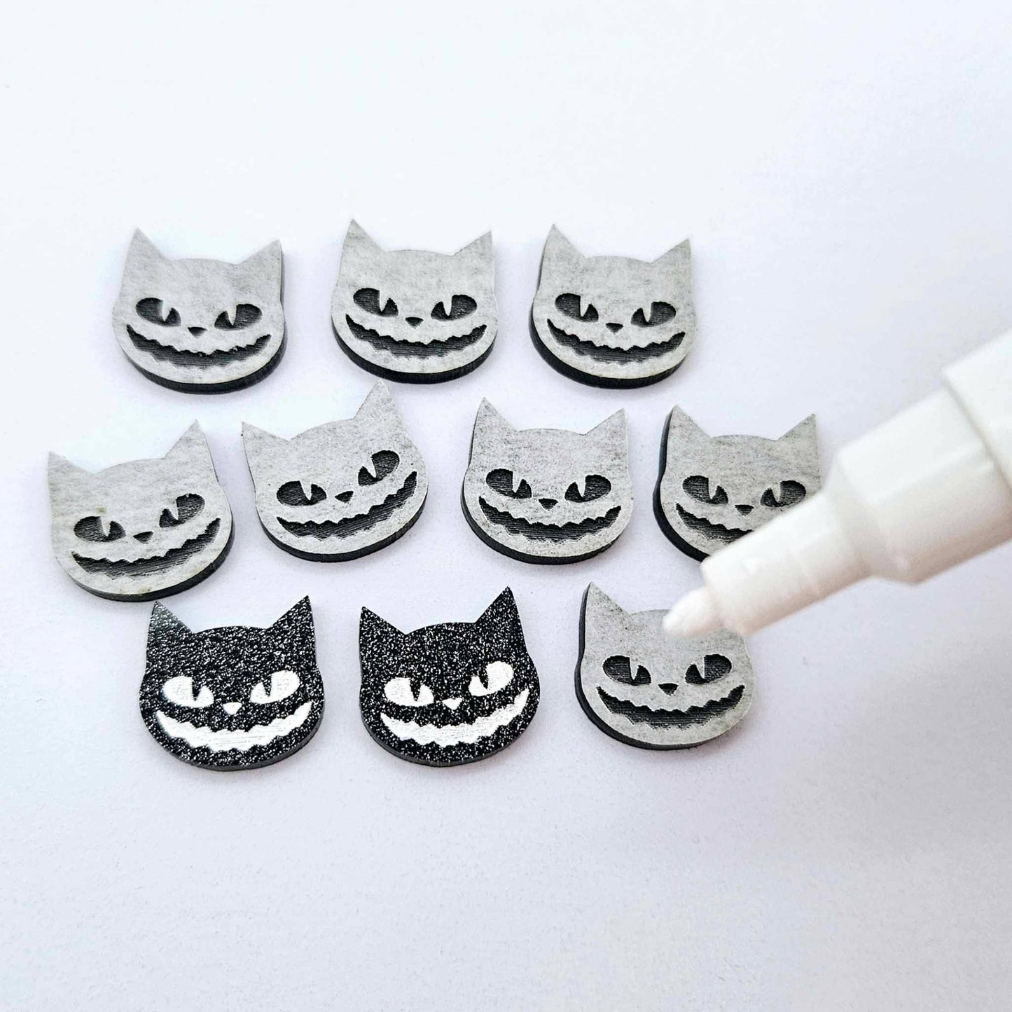 One pair of black glitter acrylic Spooky Cat stud earring blanks with white paint-filled details, surrounded by multiple unpainted pieces with protective paper on, pictured with an uncapped White Posca Paint Pen ready for paint filling