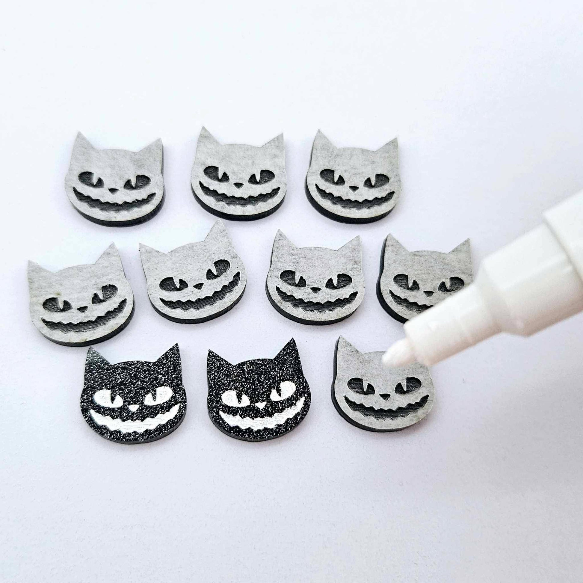 One pair of black glitter acrylic Spooky Cat stud earring blanks with white paint-filled details, surrounded by multiple unpainted pieces with protective paper on, pictured with an uncapped White Posca Paint Pen ready for paint filling