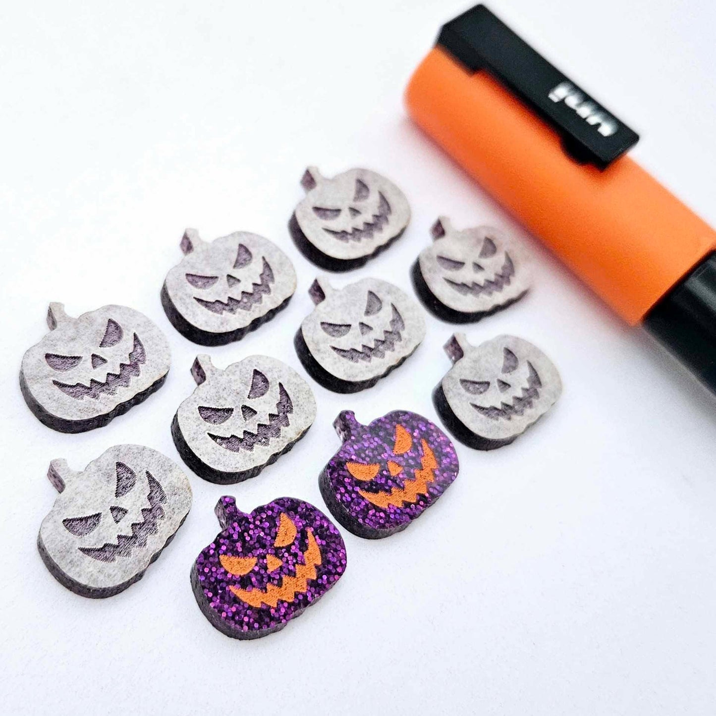 One pair of holographic purple glitter Jack-O’-Lantern stud earring blanks with orange paint-filled details, surrounded by multiple unpainted pieces with protective paper on, pictured with an Orange Posca Paint Pen