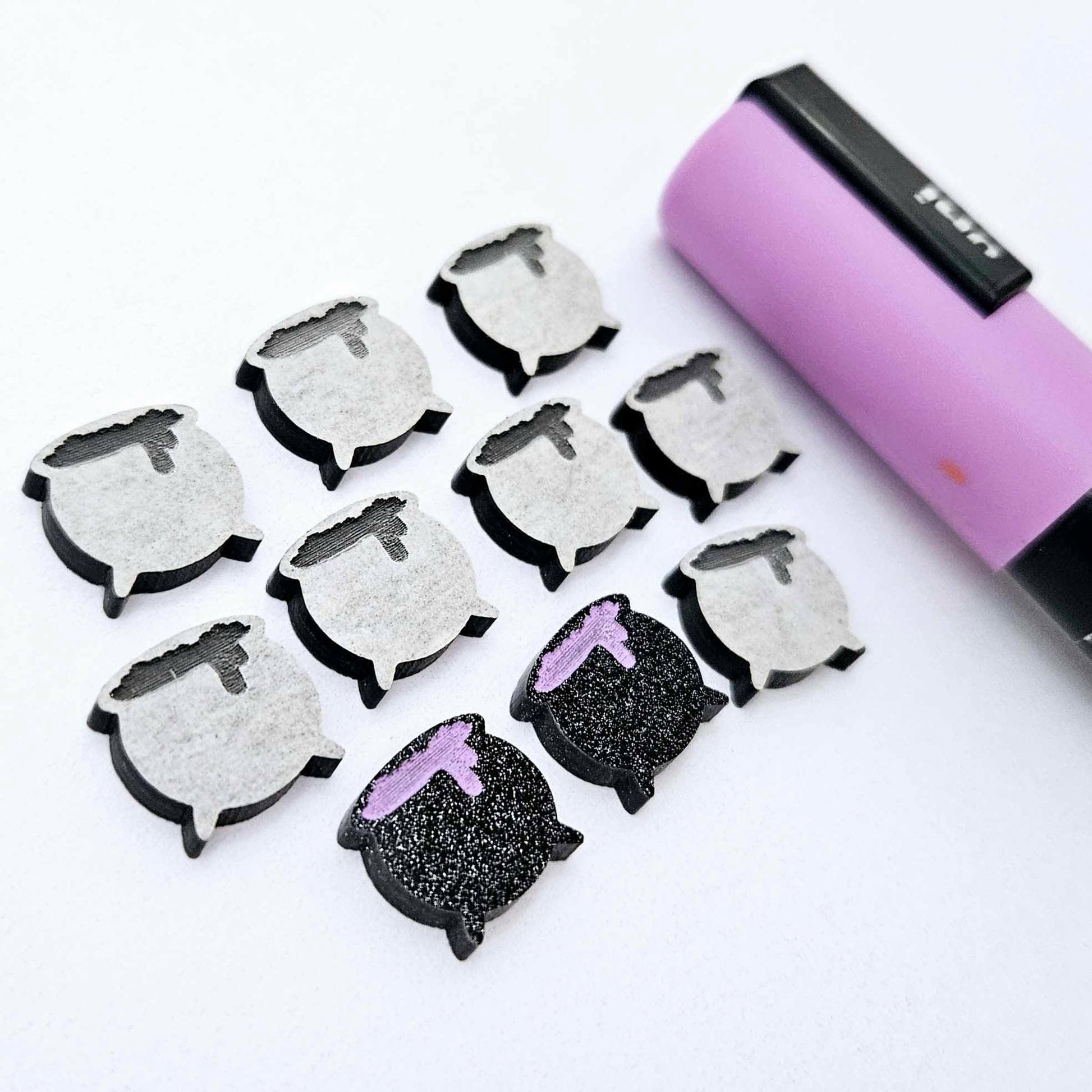 One pair of black glitter acrylic Witch's Cauldron stud earring blanks with lavender paint-filled details, surrounded by multiple unpainted pieces with protective paper on, pictured with a Lavender Posca Paint Pen.